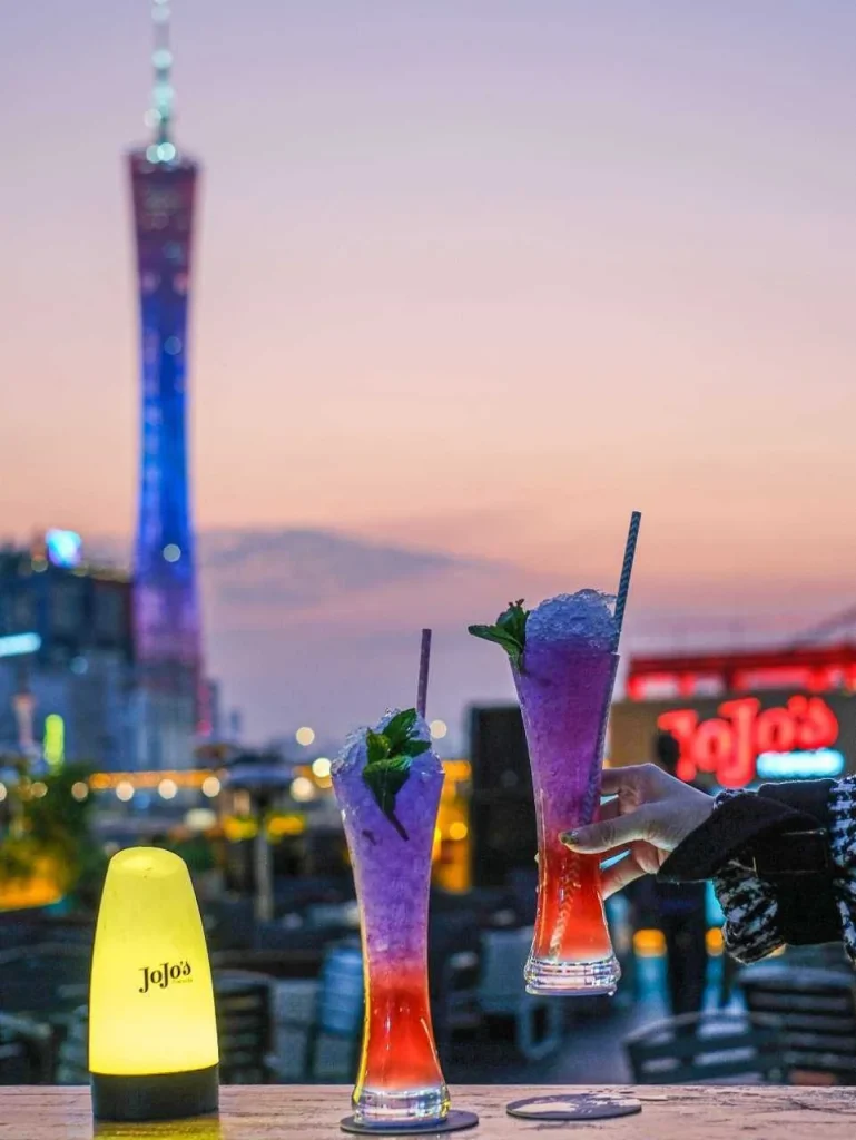 黄昏时分露台上的两杯缤纷鸡尾酒，点缀薄荷与条纹吸管，身后是广州塔